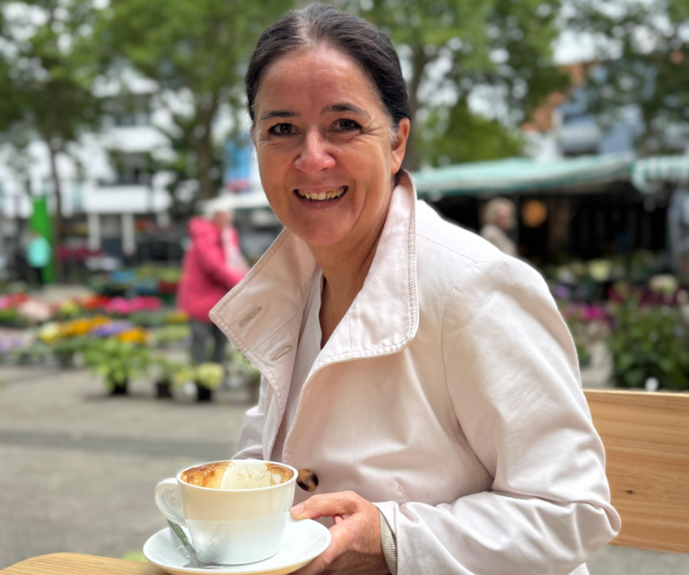 Jacqueline Turowsky privat mit Kaffee in einem Freiluftcafé, umgeben von Blumen und Bäumen und die lachend in die Kamera schaut.