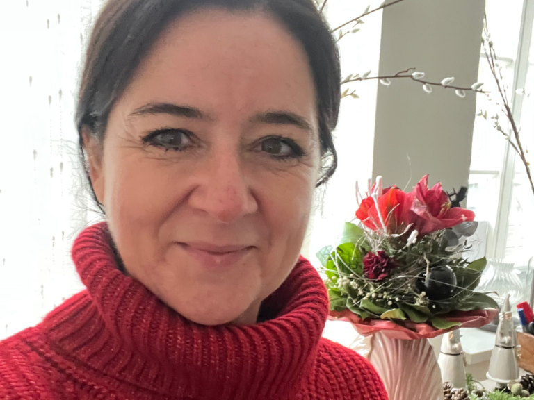 Jacqueline Turowsky privat mit dunklen Haaren trägt einen roten Pullover, im Hintergrund ein Blumenarrangement.