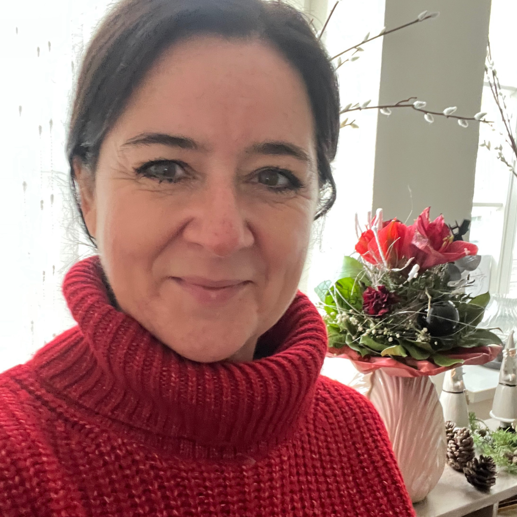 Jacqueline Turowsky privat mit dunklen Haaren trägt einen roten Pullover, im Hintergrund ein Blumenarrangement.