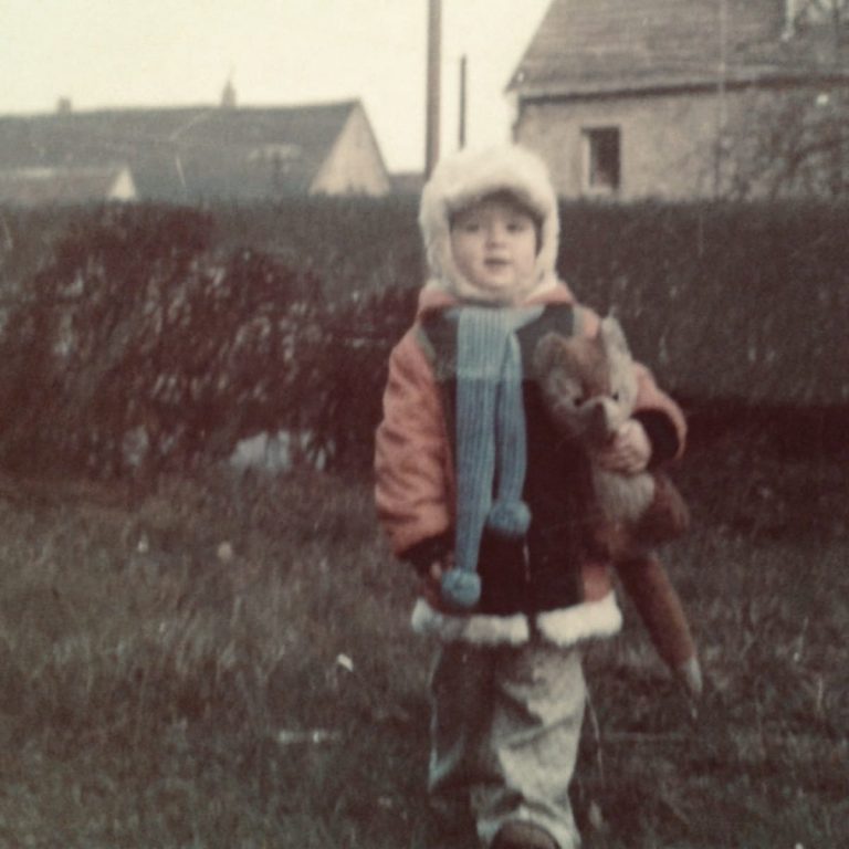 Ein Kind in warmer Kleidung hält ein Stofftier in der Hand, umgeben von Natur.